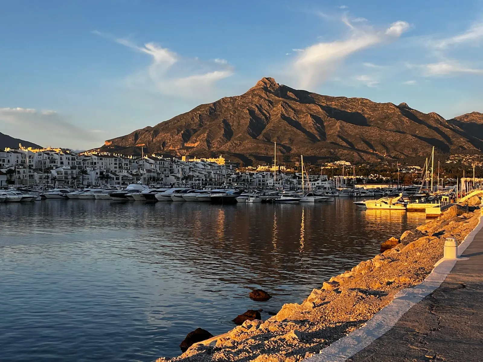 A view of the yacht harbor and white apartment buildings at the foot of the mountains in Spain at sunset - the perfect place for a house or apartment in Spain for sale with sea views.